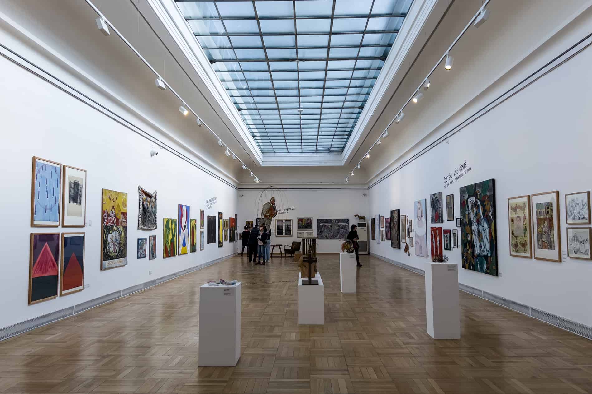 People browsing the paintings inside a modern art museum with whit walls and a glass ceiling. 