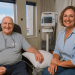 Peter, a smiling elderly man undergoing cancer treatment, sits in a hospital chair with a supportive female companion beside him, surrounded by medical equipment.