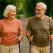 An elderly couple with light skin smiling and walking outdoors in a sunny park while holding Nordic walking poles, enjoying a healthy and active lifestyle together.