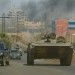 A high-resolution photograph captures a military vehicle moving through a dusty urban area during a conflict, with smoke rising in the background, surrounded by buildings and utility poles under a clear sky.