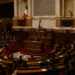 A parliamentary chamber filled with lawmakers seated in rows of red benches, listening to a speaker at the central podium under French flags during a formal political debate.