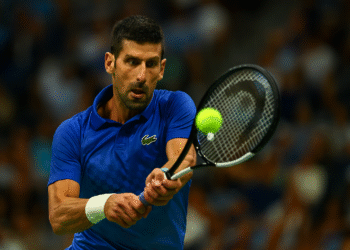 Novak Djokovic in action during a Grand Slam match, hitting a forehand shot with focus and intensity, crowd visible in the background.