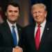 Charlie Kirk and former U.S. President Donald Trump stand side by side, smiling and shaking hands in front of a blurred American flag under warm studio lighting.
